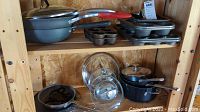 Cast iron muffin bakers, Calphalon skillet, metal baking pans, glass lids, and a cast iron pot with glass lid arranged on wood shelves.
