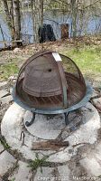 Fire pit with rusty metal and a dome mesh screen cover on a concrete round platform outdoors near a lake.