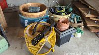 Photo showing various garden items including yellow wicker stool, blue ceramic planter, multiple pots, garden tools, and hose with sprayer.