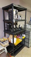 Full view of black plastic shelving unit with items on shelves for sizing reference. Shelving unit has four shelves and measures approximately 34x26x72 inches.