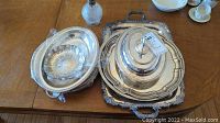 Top view of several silver plated platters and trays including covered serving dish on a rectangular ornate tray, oval platter, and a glass oil lamp without shade.