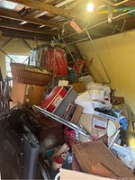 Shed interior with piled up boxes and containers including a red cooler, wicker baskets, black case, and metal pipes.