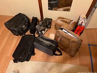 Seven assorted bags on wooden floor including two brown vintage suitcases, black briefcase, patterned bag, black bags with straps and cosmetic bag