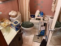 Bathroom overview showing various items on floor and counter including adult diapers, boxed safety handles, blood pressure tester, bath beauty kit, and a green toilet seat cover on the toilet.