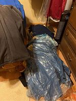 Pile of assorted denim jackets in plastic dry cleaning bags with a gray zip-up jacket and blue shirt draped over