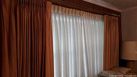 Wide view of window with soft gold pleated drapes and white sheers behind, mounted on brass rods.