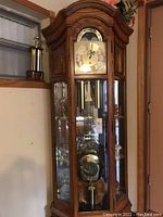 Full view of tall oak case with arched top, glass panels, brass dial and visible pendulum.