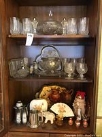 Full view of three shelves inside wooden cabinet showing crystal glassware on top and middle shelves and assorted porcelain and ceramic on bottom shelf.