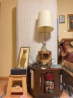 Wide view showing octagonal glass top wooden table, silver-tone ornate lamp with cream shade, vintage alarm clock, framed cat artwork, books, and other small items on a wooden stool and table near bed.