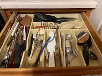 Top view of open drawer containing varied kitchen utensils including spatulas, scissors, peelers, spoons, and other kitchen tools