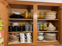 Wide view of oak cabinet filled with varied dinnerware including plates, mugs, bowls, and glassware on three shelves