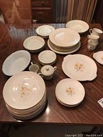Photo showing full set of dishes laid out on table, including plates, bowls, cups, sugar bowl, and serving platters with two distinct patterns: sunflower design and plain white with gold rim.
