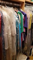View of a closet rod with multiple women's dresses and tops, some covered in plastic garment bags, including a white lace garment in front