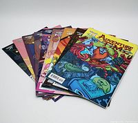 Group photo of nine Adventure Time comic books fanned out showing the front covers, showcasing colorful artwork and series numbering.
