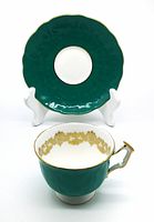 Dark teal scalloped saucer behind matching teacup showing gold floral decoration inside cup and gold trim on handle
