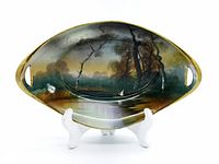 Oval Noritake serving dish with two cut-out handles, hand painted with a scenic pond and swan in flight, set on a white stand.