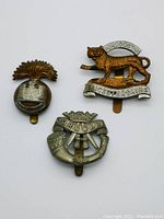 Three vintage WW2 military cap badges arranged together, showing different designs and metallic finishes.