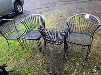 6 black metal patio chairs arranged outdoors on grass and dirt ground. Chairs have mesh seats and vertical rod fan-shaped backs.