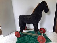 Side view of antique child's rolling horse showing black faux fur body, metal frame, red wheels, missing ears, and button eyes.