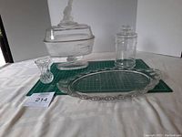Four assorted vintage or antique glass/crystal items displayed on a white cloth and green cutting mat, including a pedestal covered dish with figurine lid, a tall jar with lid, an oval serving tray, and a small vase or candle holder.