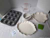 Photo showing all bake/cook ware items in the lot: Pyrex measuring cup, metal muffin pan, ceramic mortar and pestle, ceramic scalloped pie dish, ceramic leaf-shaped bowl, and green silicone finger mitt.