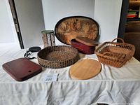 Photo showing assorted baskets, trays, cutting boards, a decorative tray, box, lantern, and wall hook arranged on a white cloth.