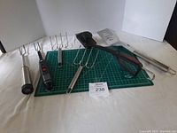 Photo of six BBQ tools arranged on a green cutting mat showing forks, brush, spatula, and tongs.