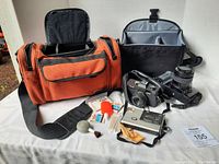Photo showing contents of lot including two canvas camera bags (one orange and one black), three cameras (Pentax with lens, vintage metal camera, compact camera), and various camera cleaning accessories.