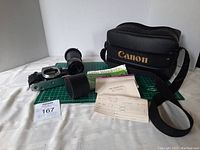 Photo showing Canon camera body, two lenses, black Canon padded camera case with strap, original paperwork, lens hood, and Lens Wonderland lens cleaning kit box on white background with green cutting mat beneath.