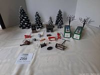 Top-down view of various Christmas village miniature accessories spread on a white cloth: 5 snow-covered pine trees, 2 boxed bare tree figurines, stone archway, cabin, sled, watering can, bench, lamppost, and other small items.