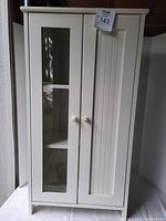 Front view of ivory wooden cabinet with two doors, one glass panel and one solid panel, both with round knobs