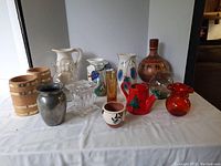 Photo showing an assortment of 15 vases and containers in different materials and sizes, arranged on a white surface in front of a plain backdrop.