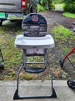 Full front view of Modela Cosco folding high chair outdoors showing the frame, floral padded cover, and tray.