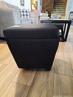 Black rolling footstool from side angle showing soft fabric covering, cushioned top, and wheels.