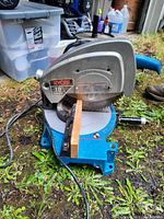 Photo showing side view of blue Ryobi 10 inch mitre saw with wooden piece clamped, on grass ground.