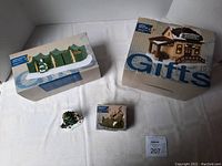 Five items shown: three boxes of village pieces and two loose figures with Christmas tree base. Boxes labeled Lighted Resident Camp Tents, Lighted Dining Hall, and Philmont Trek accessory. Loose scout and leader figures with tree present.