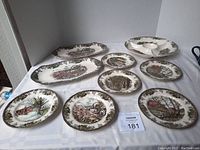 Nine-piece vintage dish and serving set displayed on a white cloth surface, showing various plates and platters with village scene designs and decorative borders.