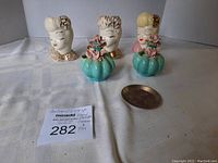 Front view of three handpainted ceramic female head figurines, two turquoise ceramic flower-shaped pieces with pink flowers, and one small metal trinket holder on white fabric background.