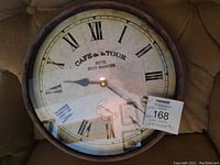 Photo of round metal wall clock with antiqued finish, Roman numeral face and inscriptions 'CAFE de la TOUR POUR BIEN MANGER'. Clock face shows some vintage crackle effect.