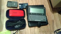 Two black battery chargers, red Samsung camera case, and electronic organizer device with screen and keyboard on a wooden surface