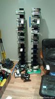 Two black DVD/CD towers filled with various DVD/CD cases. The towers are shown standing on a wooden floor next to a black chair and some electronic devices.