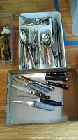 Photo of stainless steel flatware organized in a white plastic tray, along with six kitchen knives with black handles laid on cardboard.