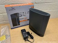 Photo showing Arris Surfboard G36 WiFi modem standing upright next to its original retail box and power adapter.