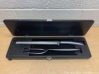 Open black storage box containing a carving knife and two-prong meat fork, both with black handles and stainless steel heads.