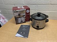 Photo showing Elite Gourmet slow cooker unit, glass lid, original box, and instruction manual on a wooden table.