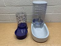 Two pet water bowls side by side against a brick wall background, one white and one purple, both with clear water reservoirs upright.
