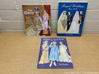 Three paper doll books related to royal weddings on a table against a cinder block wall.
