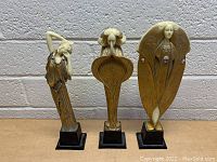 Three Art Nouveau style statues of female figures, beige color, black bases, arranged in a row against a white textured wall.