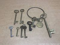 Lot of 8 vintage keys of various sizes and designs laid out on a wooden surface, including a large metal key ring holding three keys and a round tag with beehive design.