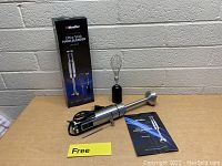 Mueller Ultra Stick Hand Blender MU-HB-02 main unit with whisk attachment, detachable blending shaft, user manual, and original box on a grey floor near a brick wall.